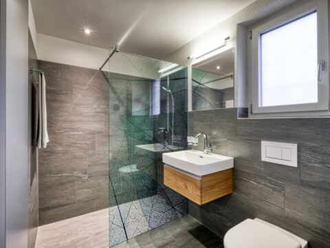 Badezimmer mit grauem Boden und grauen Wänden, einer Duschtrennwand aus Glas und einem Lavabomöbel aus Holz, Badezimmer-Umbau realisiert durch DIE SCHREINER